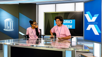 Two high school students sitting at anchor desk of a TV studio on the campus of UNC Chapel Hill.